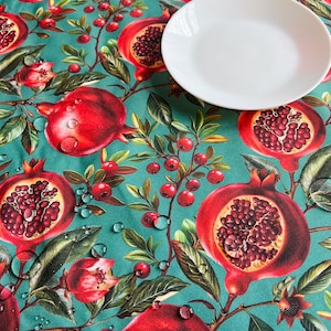 Pode incluir: Uma toalha de mesa turquesa com um padrão vibrante de romã. O design apresenta romãs grandes e maduras em tons de vermelho, com folhas verdes e pequenas bagas vermelhas. Um prato branco repousa sobre a toalha de mesa.