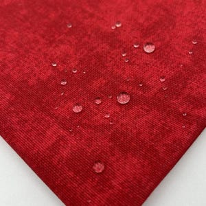 May include: Close-up of a red fabric with a textured appearance. Water droplets are visible on the surface, indicating a water-resistant or waterproof material. The fabric has a slightly mottled pattern.