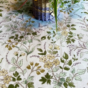 Puede incluir: Un mantel blanco con un patrón floral en tonos verdes, amarillos y morados. Una cesta de mimbre llena de un ramo de flores naranjas, amarillas y moradas se encuentra sobre el mantel.