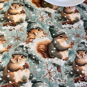May include: A light blue tablecloth featuring a repeating pattern of squirrels wearing gray knit hats, surrounded by snowflakes and pinecones. A white plate is partially visible in the image. The squirrels are holding pinecones.
