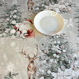 Könnte beinhalten: Festliche Weihnachtstischdecke mit einem sich wiederholenden Muster aus Weihnachtsmännern, Rentieren und geschmückten Weihnachtsbäumen. Die Farbpalette umfasst Rot, Weiß und Grün auf hellgrauem Hintergrund. Ein weißer Teller liegt auf der Tischdecke.
