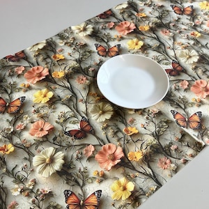 May include: A floral table runner with a butterfly and flower design. The runner features orange, yellow, and cream-colored flowers and butterflies. A white plate sits on top of the runner. The runner is on a white table.