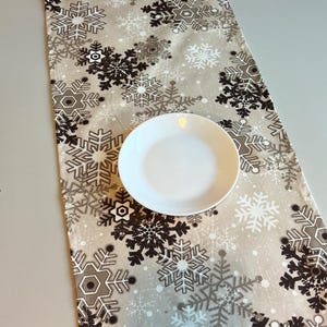 May include: A rectangular beige table runner with a repeating snowflake pattern in shades of gray and white. A white, round plate sits on top of the runner. The runner is likely for holiday or winter decor.
