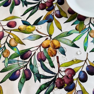 May include: A white tablecloth featuring a watercolour-style print of olive branches with green and purple olives and elongated green leaves. Several clear water droplets are visible on the fabric surface, alongside a plain white ceramic plate.