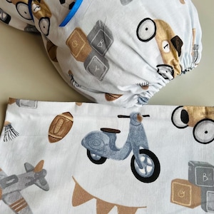 May include: A white fabric with a blue ribbon tied around it. The fabric has a pattern of various toys, including a scooter, a plane, a soccer ball, and building blocks. The fabric is folded in half and the ribbon is tied in a bow.