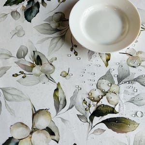 Może przedstawiać: Biały ceramiczny talerz na kwiecistym obrusie. Obrus ma powtarzający się wzór białych jagód, zielonych liści i szarych liści. Krople wody rozrzucone po powierzchni. Motywem przewodnim jest natura i posiłek.