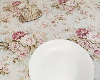 Puede incluir: Un plato de cerámica blanco con borde festoneado se encuentra sobre un mantel floral. El mantel tiene un fondo azul claro con flores rosas y blancas.
