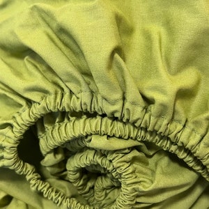 以下が含まれることがあります： 緑色のフィットシーツのアップ画像。シーツは柔らかく滑らかな生地でできており、マットレスにぴったりとフィットするように設計されています。