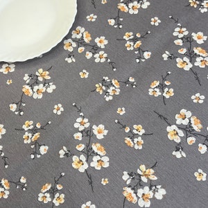 以下が含まれることがあります： 白と黄色の花柄のグレーのテーブルクロス。テーブルクロスは微妙な質感と、小さな白い花に黄色の芯がついた繰り返し模様があります。