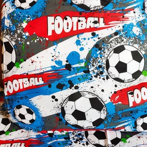 May include: White fabric with a blue, red, and gray splatter paint design. The fabric features black and white soccer balls and the word "Football" repeated throughout.