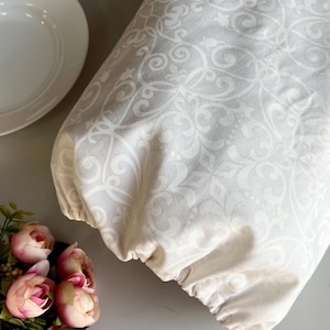 May include: A cream-coloured fabric cover with a white scroll pattern, possibly for a kitchen appliance. A white plate and pink roses are in the background. The fabric has a repeating scroll pattern.