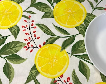 May include: A white tablecloth with a vibrant lemon and leaf pattern. The design features bright yellow lemon slices, green leaves, and small red berries. A white plate is partially visible on the right side, suggesting a dining setting.