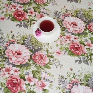 Könnte beinhalten: Eine weiße Teetasse und Untertasse mit Tee, auf einer floralen Tischdecke. Die Tischdecke zeigt ein Muster aus rosa und roten Rosen, grünen Blättern und grauen Akzenten. Ein kleines rosa Dekorationsobjekt befindet sich in der Nähe der Tasse.