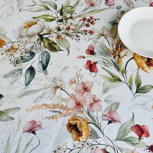 May include: White floral tablecloth with a watercolor design featuring pink, red, yellow, and blue flowers, green leaves, and water droplets. A white plate is in the upper right corner. The tablecloth is suitable for a dining table.