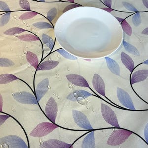 Op de afbeelding: Een wit tafelkleed met een bladmotief in paars en blauw. Waterdruppels zijn verspreid over het oppervlak. Een wit bord is in het midden van het tafelkleed geplaatst.