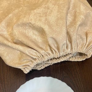 May include: A beige fabric cover with a gathered elastic edge. The cover is on a wooden surface next to a white ceramic plate.