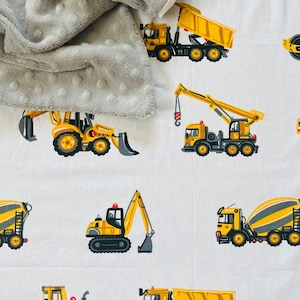 May include: A white fabric with a repeating pattern of yellow construction vehicles. The vehicles include a cement mixer, a dump truck, an excavator, a crane, and a road roller. A gray textured blanket is partially visible in the top left corner of the image.