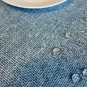 Op de afbeelding: Een close-up van een blauwe tafelkleed met een geweven patroon. Waterdruppels zijn verspreid over het oppervlak.