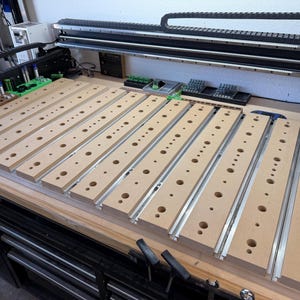 May include: A CNC machine bed featuring a series of parallel light brown MDF slats with pre-drilled circular holes of varying sizes. The slats are mounted on a metal frame with integrated silver aluminum T-slot tracks for securing workpieces.