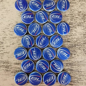 May include: A collection of 25 blue and white Bud Light bottle caps. The caps are arranged in a loose circle on a light brown wooden surface.