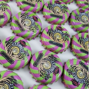 May include: A collection of 15 bottle caps from Three Floyd's Brewing Co. The bottle caps are green and pink with a white logo and text. The logo features two hands holding a circle with the letters "FFF" inside. The text reads "Three Floyd's Brewing Co."