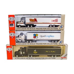 May include: Three boxed HO scale model semi-trucks stacked vertically. The top truck is white with a horse-drawn carriage graphic. The middle truck is white with a QuadGraphics logo. The bottom truck is black with a Hawkeye logo and text.