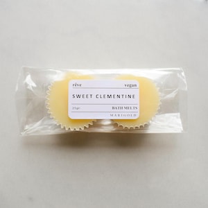 May include: Two round, yellow bath melts in a clear plastic bag. The label reads "r&ecirc;ve vegan SWEET CLEMENTINE BATH MELTS MARIGOLD 25g"