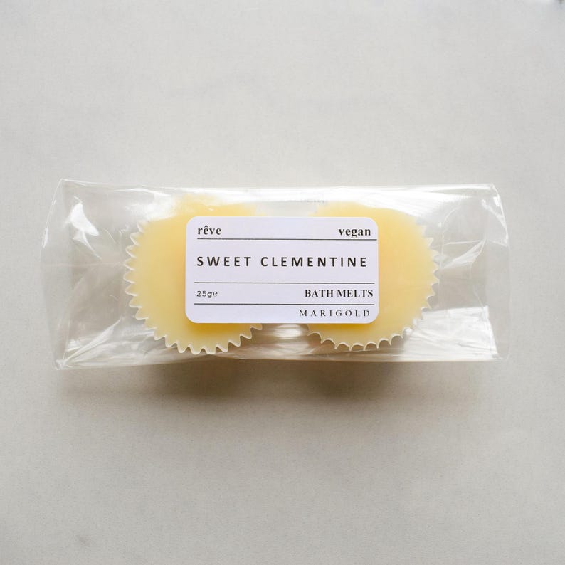 May include: Two round, yellow bath melts in a clear plastic bag. The label reads "r&ecirc;ve vegan SWEET CLEMENTINE BATH MELTS MARIGOLD 25g"