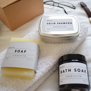 May include: A set of bath and body products in a Siberian Fir scent. The set includes a bar of soap, a tin of solid shampoo, and a jar of bath soak. All products are labeled with the text "Man Kit" and "Siberian Fir".