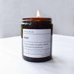 Peut inclure: Un bocal en verre brun contenant une bougie avec une étiquette blanche sur laquelle est inscrit "Genius No.01 REST let your body rest with a blend of lavender & patchouli 30 hrs 100% plant wax infused with essential oils MARIGOLD". La bougie est allumée et la flamme est visible.