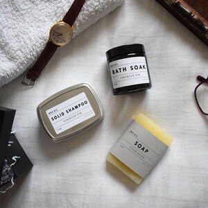 May include: A set of three bath products in a Siberian Fir scent. The products include a solid shampoo bar in a silver tin, a bar of soap, and a bath soak in a black jar.