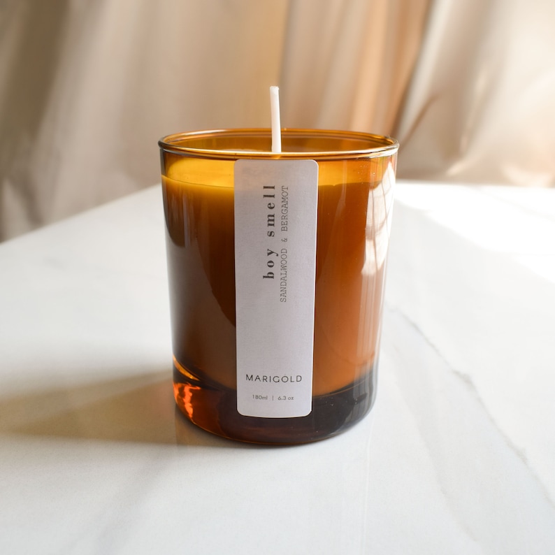May include: A brown glass candle jar with a white label that reads "boy smell SANDALWOOD & BERGAMOT" and "MARIGOLD 180ml | 6.3 oz".