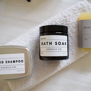 May include: A set of three bath products in a Siberian Fir scent. The products include a black jar labeled "Bath Soak", a silver tin labeled "Solid Shampoo", and a bar of soap labeled "Soap".