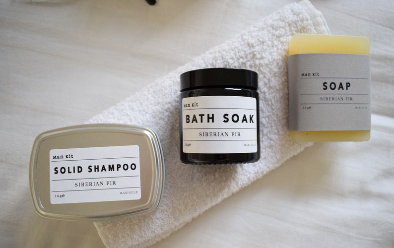 May include: A set of three bath products in a Siberian Fir scent. The products include a black jar labeled "Bath Soak", a silver tin labeled "Solid Shampoo", and a bar of soap labeled "Soap".