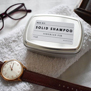 May include: A silver tin with a white label that reads "Man kit SOLID SHAMPOO Siberian Fir MARIGOLD 30g e". The tin is sitting on a white towel.