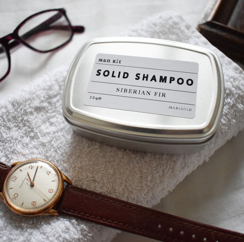 May include: A silver tin with a white label that reads "Man kit SOLID SHAMPOO Siberian Fir MARIGOLD 30g e". The tin is sitting on a white towel.