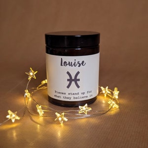 May include: A brown glass candle jar with a white label. The label says "Louise" and features the Pisces astrological symbol. The text on the label reads "Pisces stand up for what they believe in". The candle jar is surrounded by a string of white lights shaped like stars.