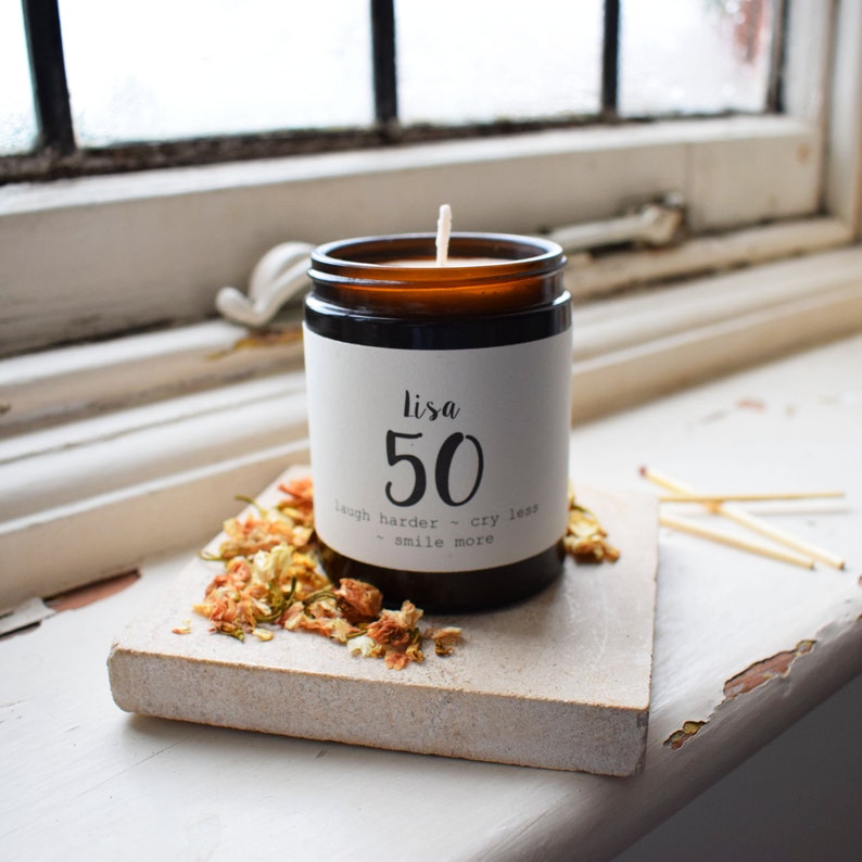 Puede incluir: Una vela de vidrio marr&oacute;n con una etiqueta blanca que dice "Lisa 50 laugh harder - cry less - smile more". La vela est&aacute; sentada sobre una superficie blanca con p&eacute;talos de flores secas.