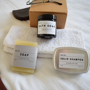May include: A set of three bath and body products in a white towel. The products are a black jar labeled "Bath Soak Siberian Fir", a white bar of soap labeled "Soap Siberian Fir", and a silver tin labeled "Solid Shampoo Siberian Fir".