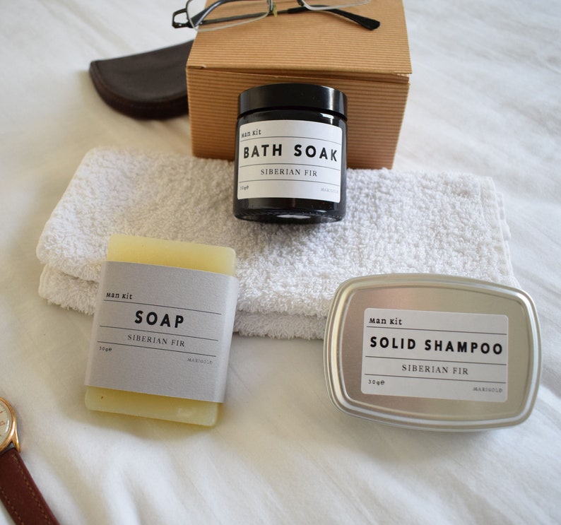 May include: A set of three bath and body products in a white towel. The products are a black jar labeled "Bath Soak Siberian Fir", a white bar of soap labeled "Soap Siberian Fir", and a silver tin labeled "Solid Shampoo Siberian Fir".