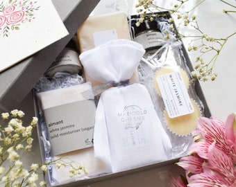 May include: A gift box filled with bath and body products, including a white jasmine scented candle, a white jasmine solid moisturizer, a white jasmine bath melt, and a white muslin bag with a MARIGOLD CHARMS label.