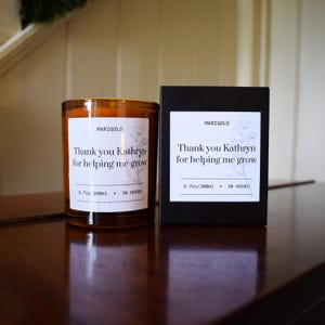 Könnte beinhalten: Ein bernsteinfarbenes Kerzenglas und eine schwarze Schachtel, beide mit einem weißen Etikett mit dem Text MARIGOLD, Thank you Kathryn for helping me grow, 6.7oz/200ml, 30 HOURS und einer feinen Strichzeichnung eines Blumenstängels versehen.