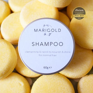 May include: A white round container with a silver lid. The container has the text "MARIGOLD SHAMPOO" printed on it. The text "clementine & neroli to nourish & shine for normal hair" is printed below the product name. The container also has the text "60g e" printed on it. The container is surrounded by yellow solid shampoo bars.