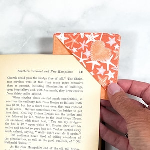 May include: A close-up of a handmade fabric bookmark with a peach background and a white heart with a stitched outline. The bookmark is decorated with white and orange starfish and stars. The bookmark is being used to mark a page in a book about Southern Vermont and New Hampshire.
