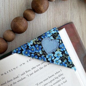 May include: A blue and white fabric bookmark with a heart-shaped design. The bookmark is folded into a triangle shape and is placed on top of an open book. The book is titled "The Writing on the Wall".