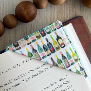 May include: A white fabric bookmark with a pattern of colorful wine bottles. The bookmark is folded into a triangle shape and is placed on a book with the text "THE WRITING ON THE" visible.