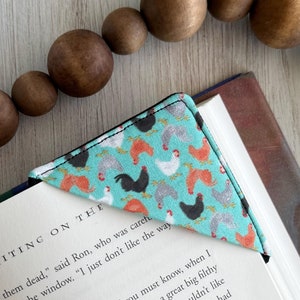May include: A turquoise blue fabric bookmark with a pattern of black, white, and orange chickens. The bookmark is folded into a triangle shape and is placed on a book with the text "THE WRITING ON THE" visible.