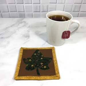 May include: A brown quilted coaster with a green leaf design and a yellow border. A white mug with a tea bag that says "Chai Spice Black Tea" is sitting on the coaster.