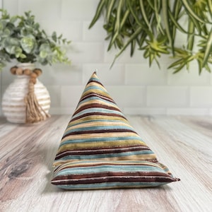 May include: A triangular pillow with horizontal stripes in shades of brown, teal, and tan. The pillow is made of fabric and sits on a wooden surface. The background includes a white brick wall and greenery.