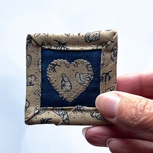 May include: A small, square fabric patch with a navy blue center and a tan border. The border is patterned with black botanical and seashell designs. A heart-shaped applique with a seashell design is centered on the navy blue fabric.